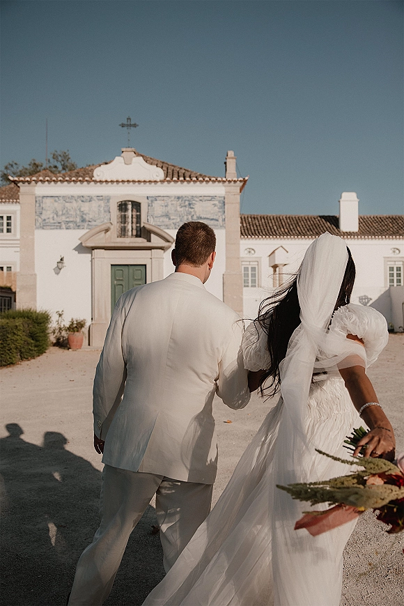 Couple portrait of bride and groom walking away, holding hands as her long veil trails, bouquet in hand before a historic tile mural facade