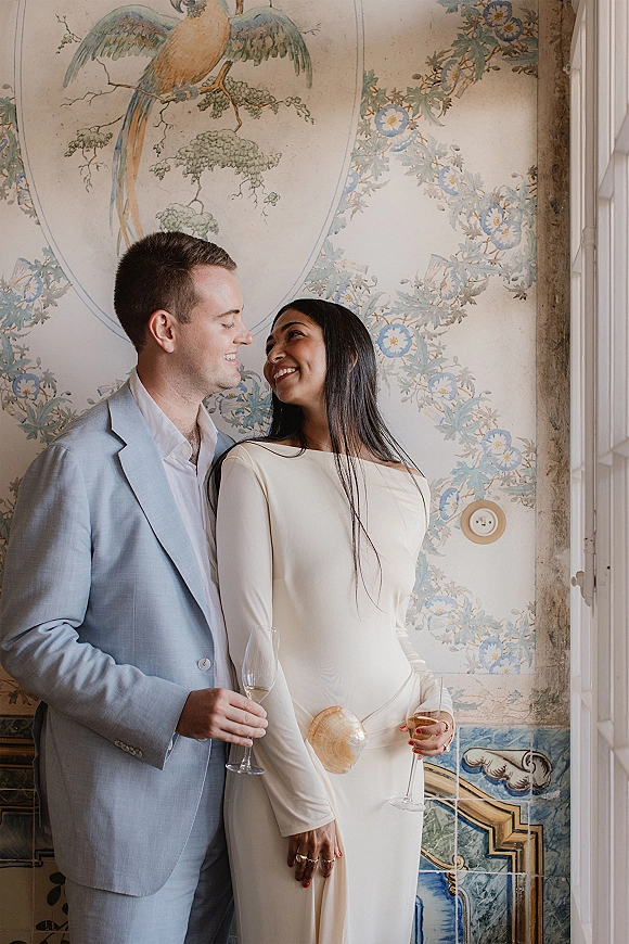 Couple portrait of bride and groom smiling, holding champagne flutes in window light against floral wallpaper and bird mural backdrop