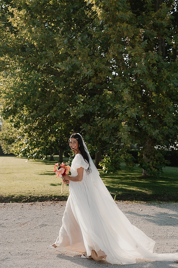 Bridal portrait of a bride looking back while walking on a sunlit gravel path, holding a coral bouquet with a long veil flowing behind