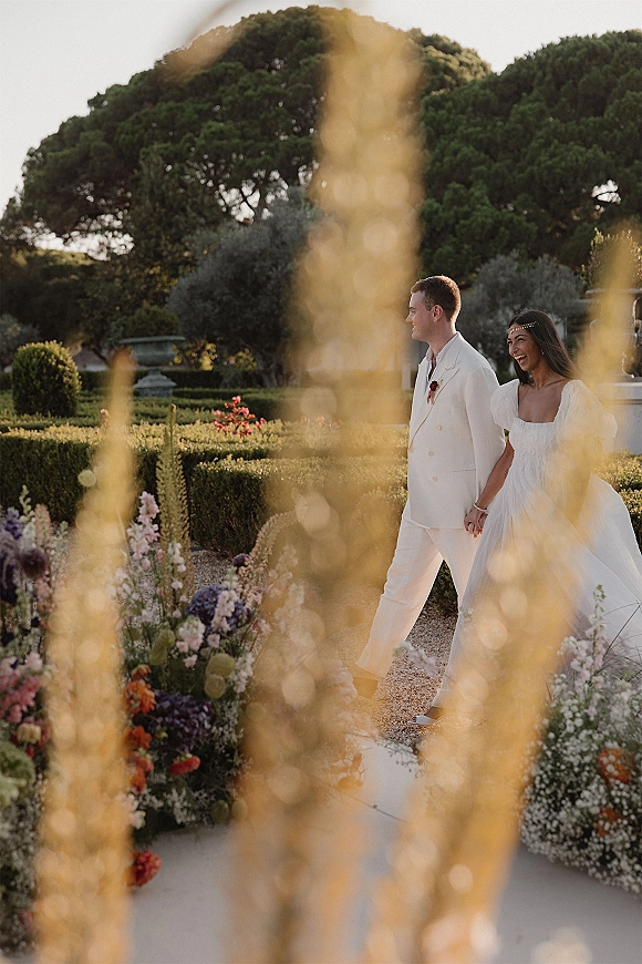 Wedding couple portrait of bride in a strapless gown and groom in a white suit holding hands, walking down a hedge-lined garden path with florals