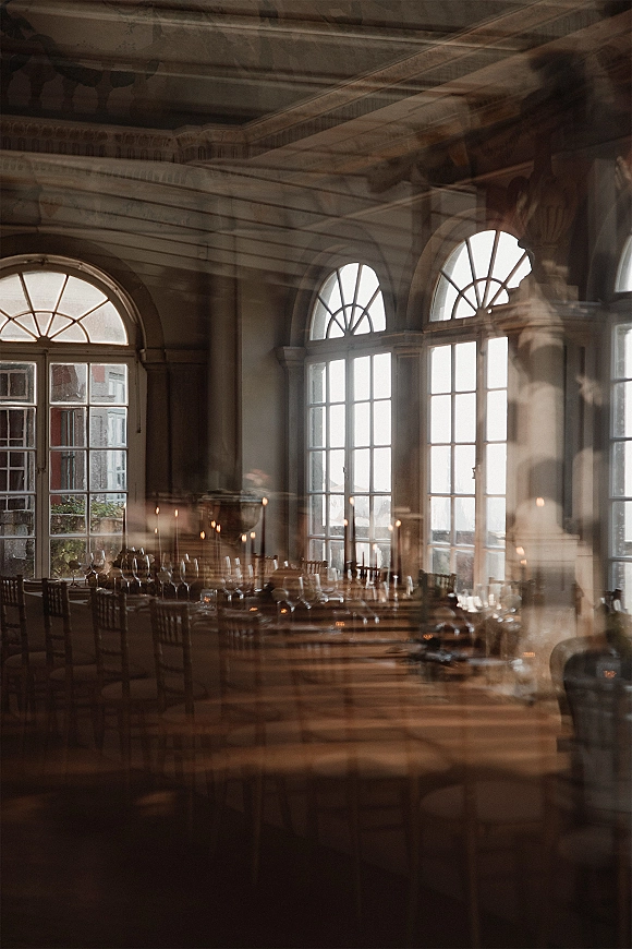 Reception tablescape with taper candles in holders, wine glasses and place settings on wooden tables beneath arched windows in natural light