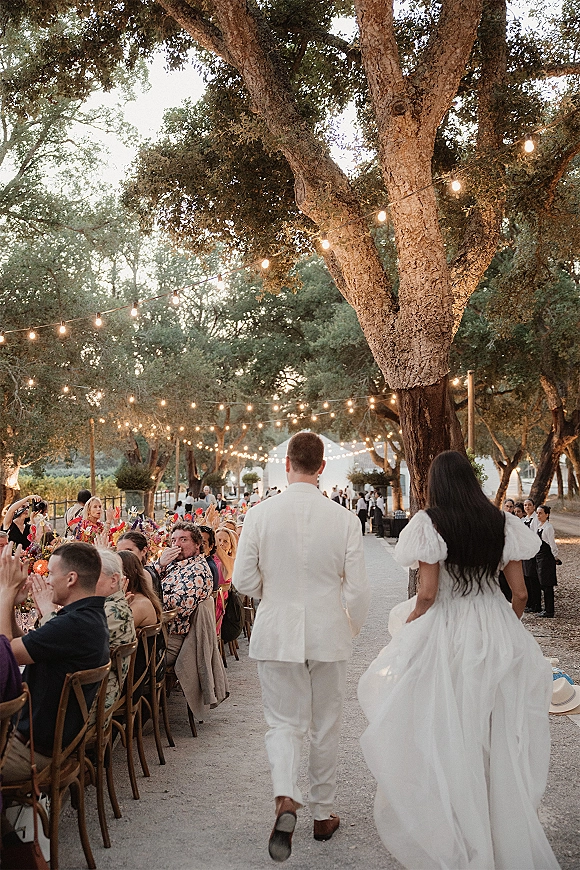 Wedding reception entrance as newlyweds walk into reception past long banquet tables with string lights, candles, and cheering guests under trees at dusk