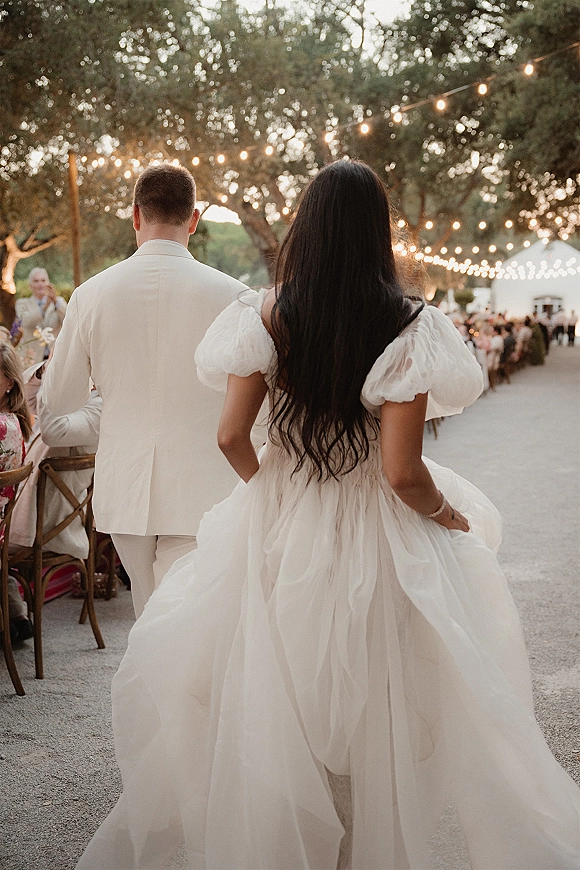 Wedding recessional as bride and groom walk away on a gravel path, her puff-sleeve tulle gown under string lights by a banquet table