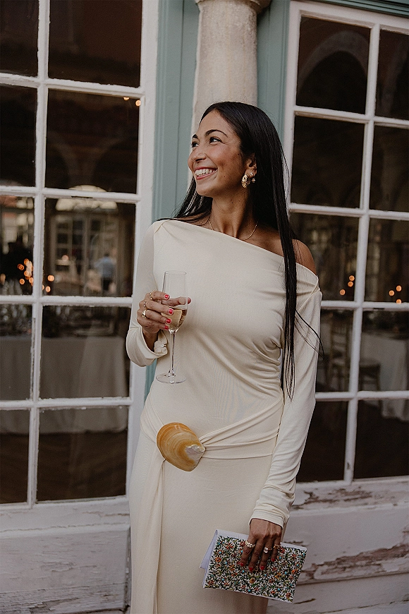 Wedding guest outfit in a cream one shoulder dress holding a champagne flute, red manicure and gold earrings by windowed doorway