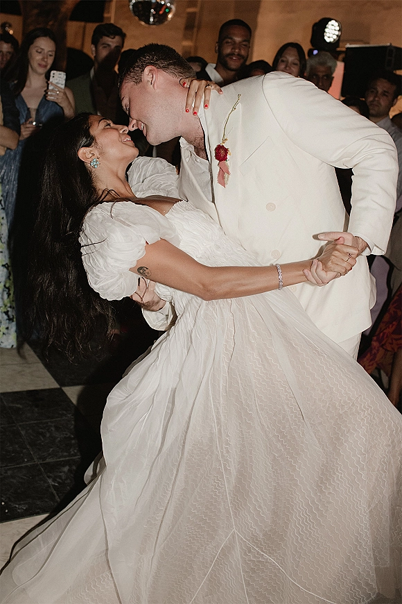 First dance dip as groom in a white suit lowers bride in an off-the-shoulder wedding dress under a disco ball, guests watching nearby