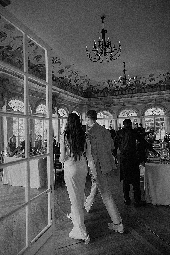 Reception entrance as newlyweds entering reception holding hands, bride in long sleeve dress and groom in suit beneath chandeliers and arched windows