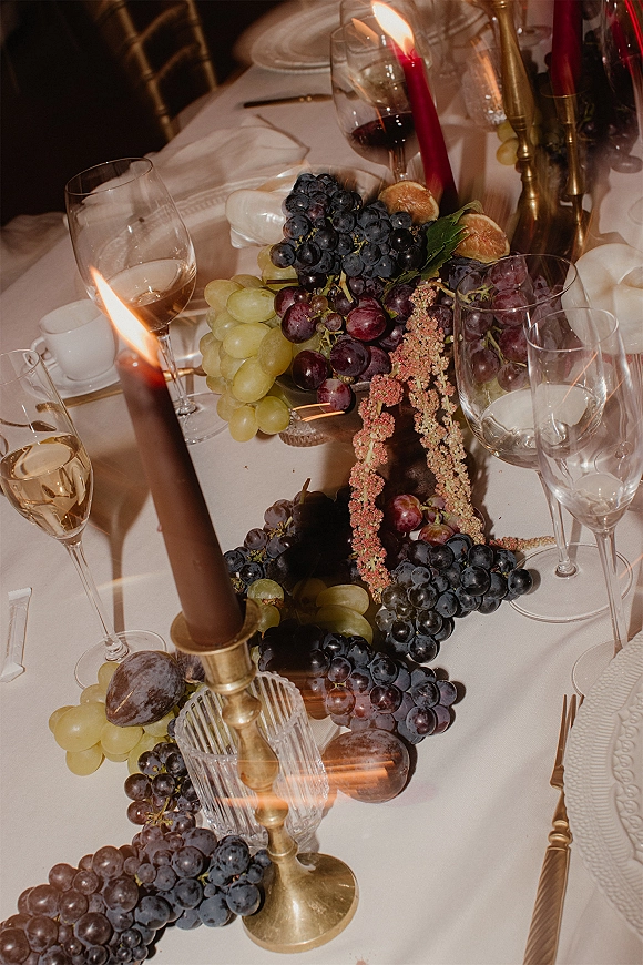 Reception tablescape with a wedding table centerpiece of grapes and figs, taper candles in brass candlesticks, wine glasses on white linens