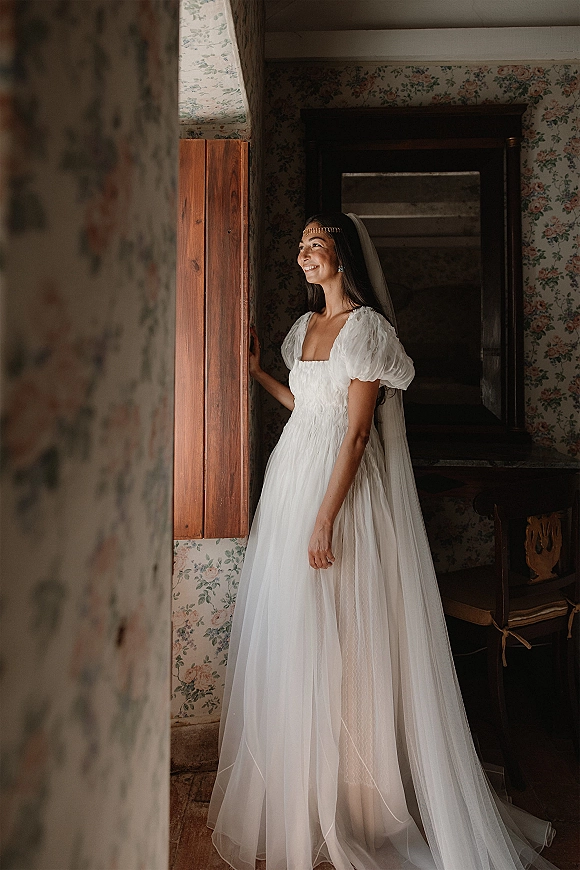 Bridal portrait of a bride standing by window in a puff sleeve wedding dress and veil, natural light on floral wallpaper backdrop