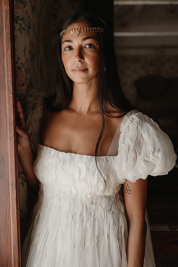 Bridal portrait of a bride with veil in a white puff sleeve wedding dress, standing in a doorway with floral wallpaper behind her.