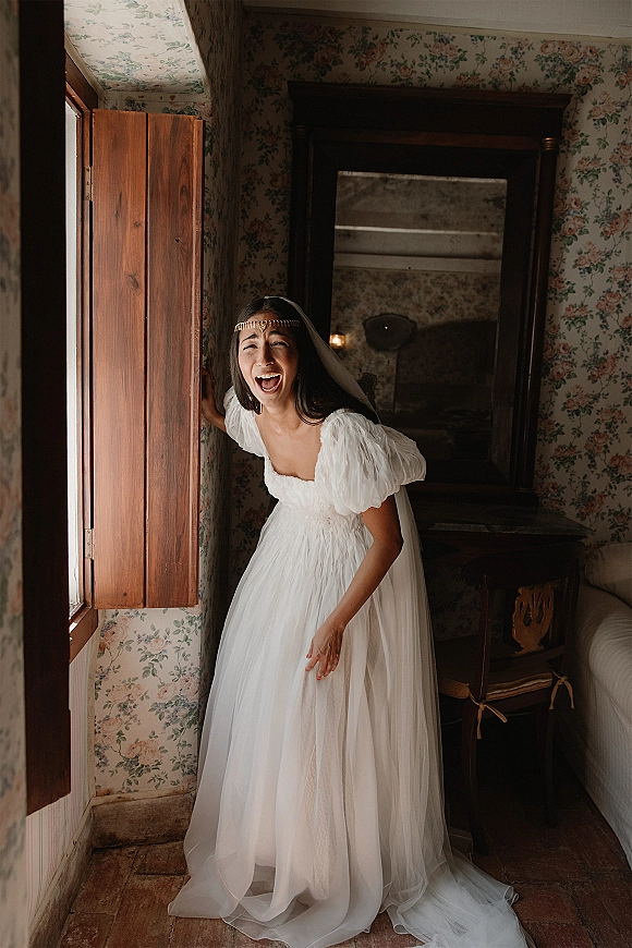 Bridal portrait of a laughing bride in a puff sleeve wedding dress and veil by a window with wooden shutters in a floral room