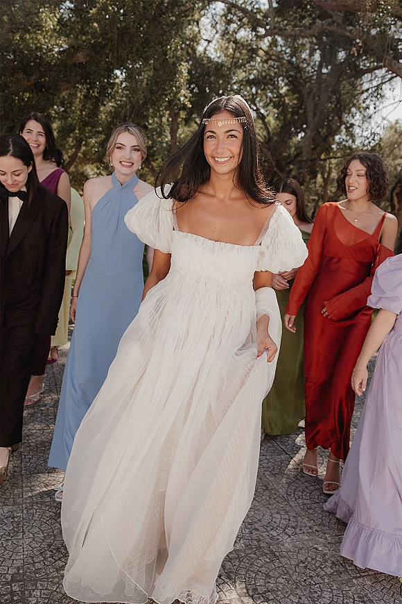 Bride portrait in an off the shoulder wedding dress with puff sleeves and gold forehead headpiece, walking a garden path with bridal party behind
