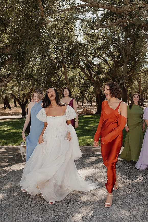Bride with bridesmaids walking and laughing in mismatched dresses beside oak trees, bride in a puff sleeve gown on a sunlit walkway