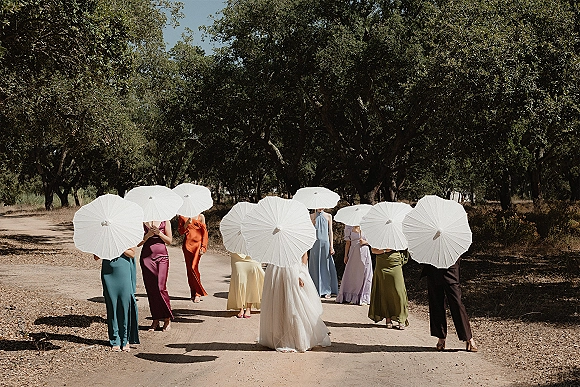 Bridesmaid group portrait with white parasol bridesmaids in colorful dresses beside the bride on a dirt road under oak trees