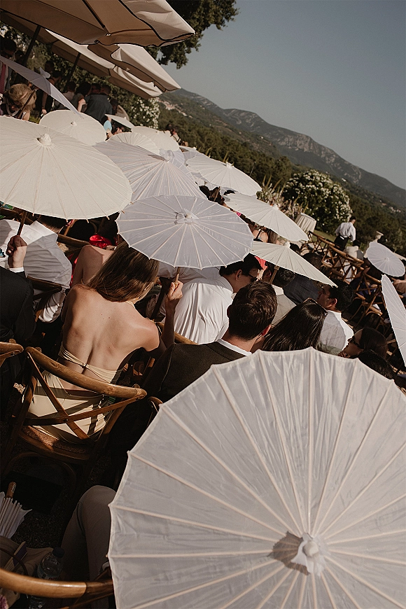 Outdoor wedding ceremony with wedding guest parasols shading wooden folding chairs on a lawn, set against mountains, trees, and clear sky