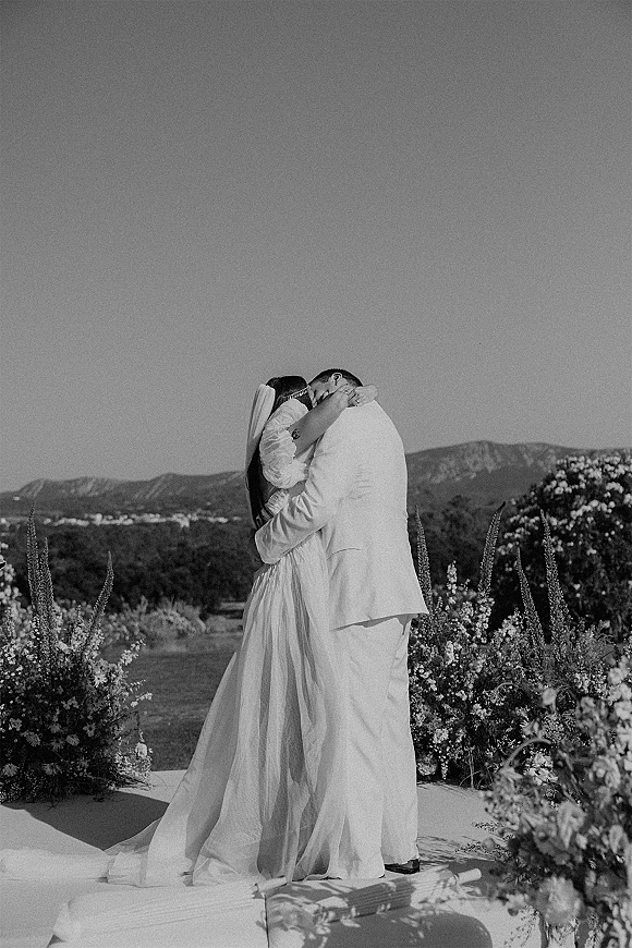 Wedding kiss as bride in long sleeve dress and veil embraces groom in white suit on aisle runner with mountains and open sky behind