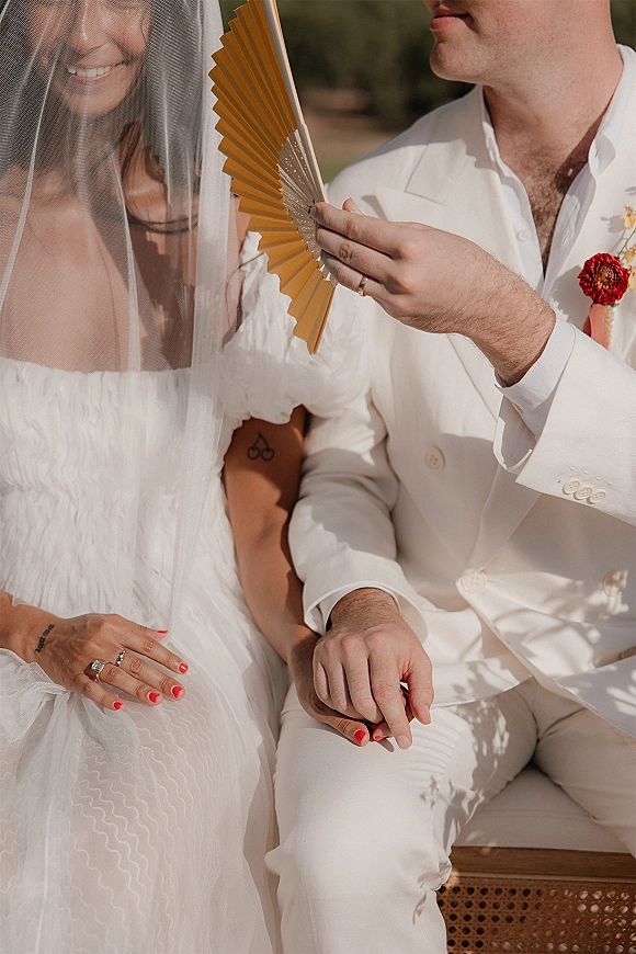 Couple portrait of bride and groom holding hands on a bench, her sheer veil over an off-shoulder gown in sunlit greenery shadows