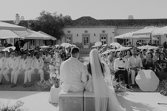 Wedding ceremony with bride and groom seated from behind, bride’s long veil trailing as guests hold parasols in a villa courtyard