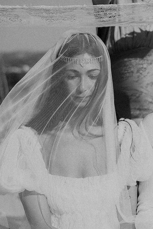 Bridal portrait of a bride under veil, looking down in a puff-sleeve gown with lace trim and forehead jewelry against a distant landscape