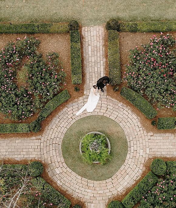 Couple portrait from above of bride and groom holding hands, her white dress train flowing on a brick walkway in a circular boxwood garden courtyard