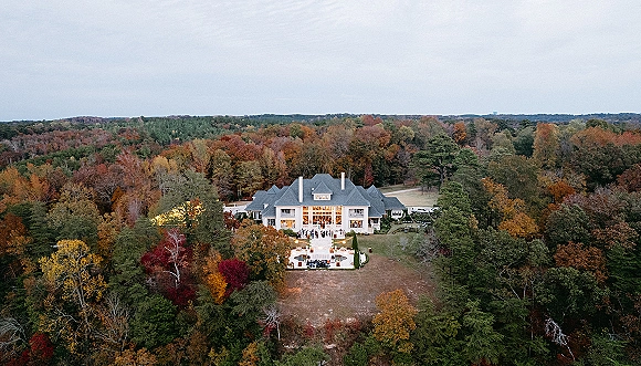 Wedding venue exterior with aerial wedding venue view, showing patio cocktail tables and outdoor lounge seating beneath string lights by fall forest lawn