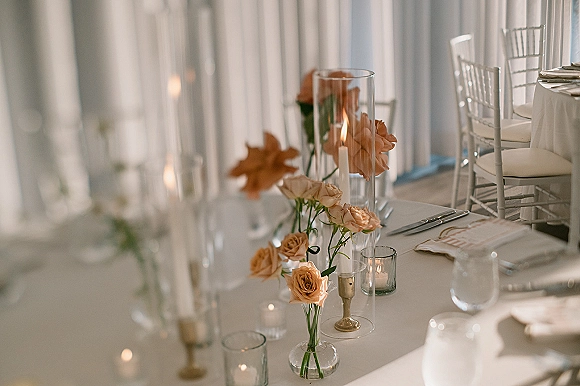 Reception tablescape with wedding table decor featuring peach rose bud vases and taper candles on a white tablecloth beneath sheer drapery