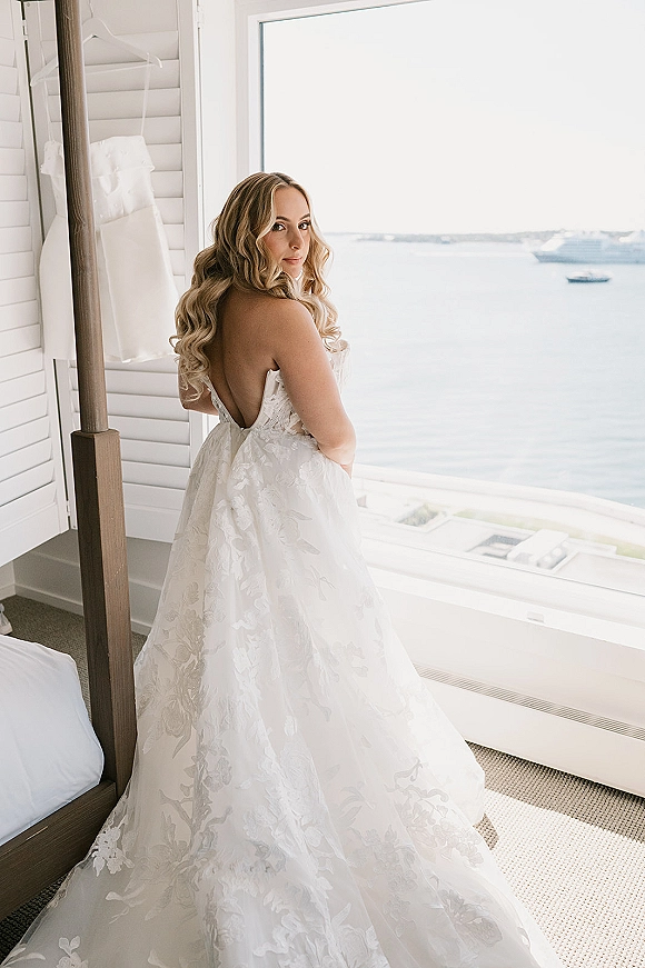Bridal portrait of bride looking over shoulder in a strapless lace wedding dress with cathedral train, by bright window and ocean view balcony