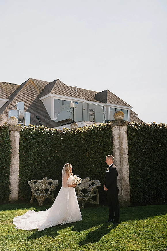Wedding first look as bride in strapless ball gown and veil approaches groom in black tuxedo, holding a white bouquet by an ivy wall