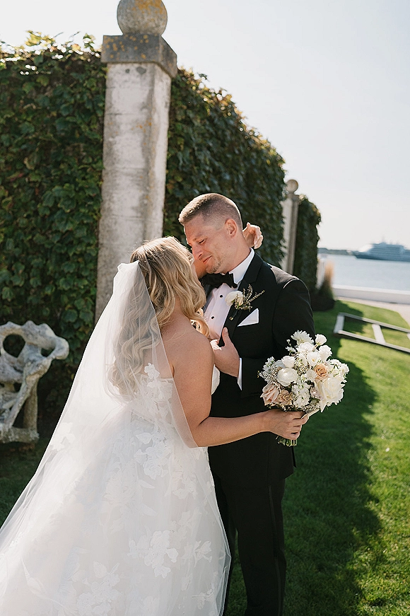 Couple portrait of bride and groom embrace, holding a wedding bouquet by an ivy wall with stone pillar and ocean beyond