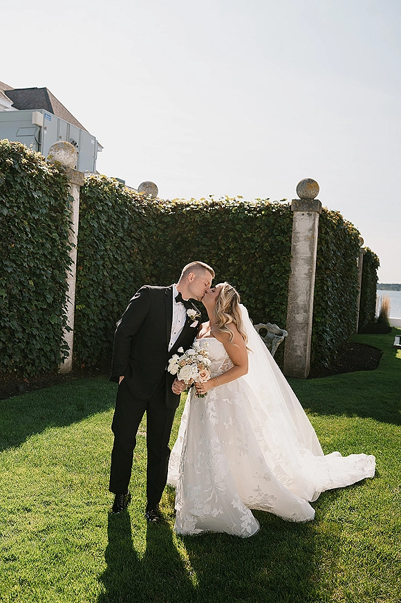 Wedding kiss portrait of bride and groom kissing, her lace dress and veil flowing as she holds a bouquet by an ivy wall and stone pillars