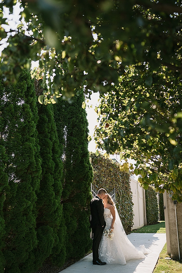 Wedding kiss portrait of bride and groom kiss, her cathedral veil and white bouquet against a sunlit vine-covered gate walkway