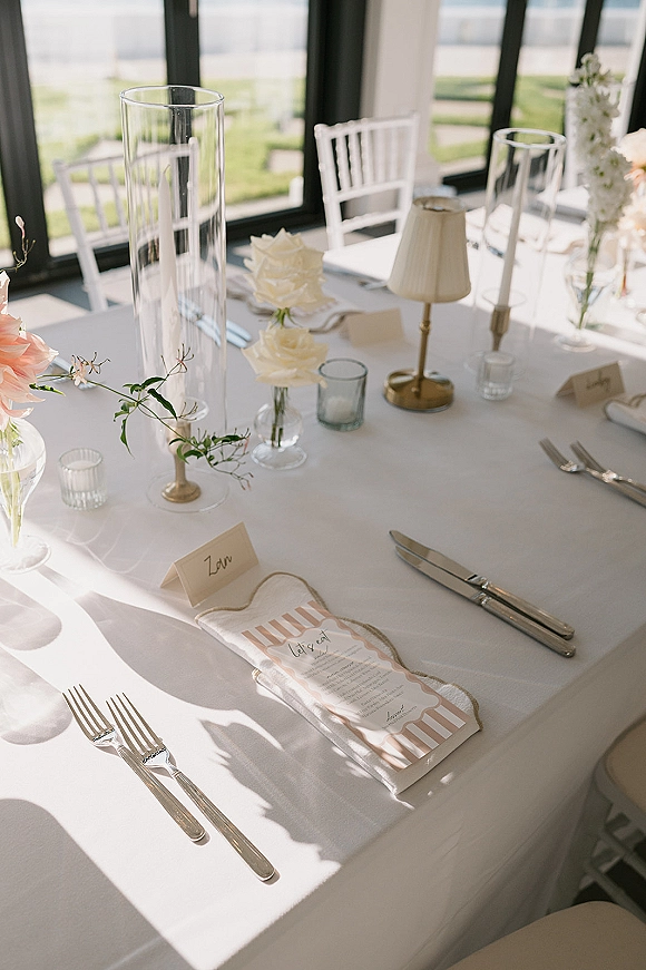 Reception tablescape with wedding place setting on white tablecloth, blush and white roses, taper candles and hurricane vases by large windows.
