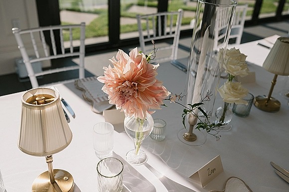 Reception tablescape with wedding table centerpiece of blush dahlias and white roses, taper candles, place cards, and window-lit lawn view