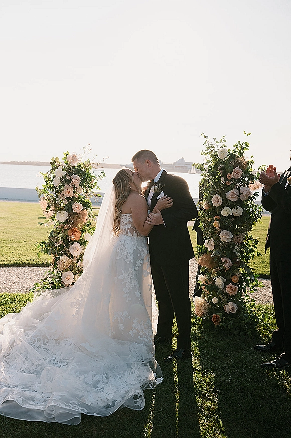 Ceremony kiss at an outdoor wedding ceremony beneath a rose wedding arch, bride in lace train and veil kissing groom in tux by the waterfront lawn