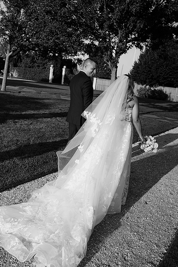 Couple portrait of bride and groom walking away on a garden gravel path, her long wedding veil trailing as he looks over his shoulder