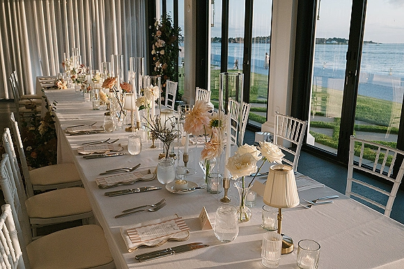 Reception tablescape with a long banquet table wedding setup, blush and white roses, taper candles, and waterfront views through tall windows