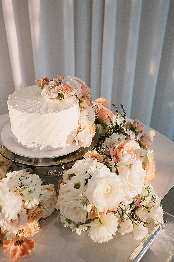 Wedding cake with white buttercream frosting topped with peach and white roses and greenery on a black stand beside a cake knife on a draped table