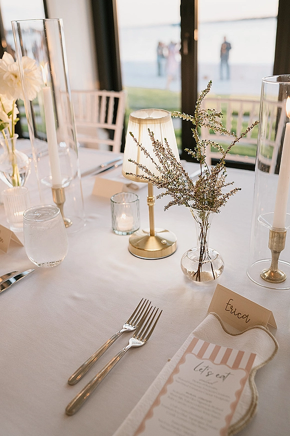 Reception tablescape with wedding table decor, taper candles in glass hurricanes, gold candlesticks and greenery by large windows overlooking water