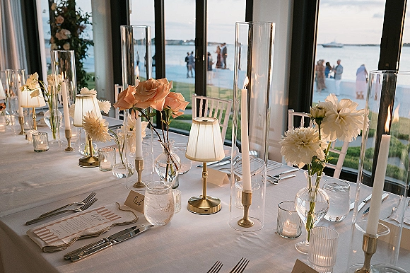 Reception tablescape with taper candle centerpiece in glass hurricanes, gold candlesticks and blush florals by floor-to-ceiling windows overlooking water