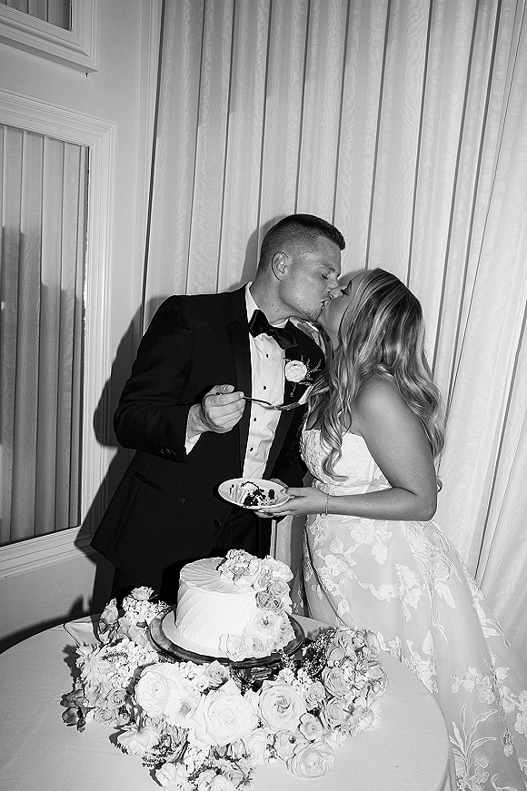 Wedding cake cutting kiss as bride in strapless lace gown and groom in black bow tie tux share a bite beside floral-topped cake table indoors
