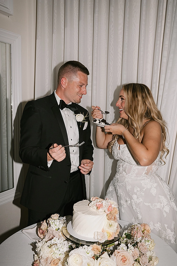 Wedding cake cutting as bride and groom feed each other a bite with forks beside a floral-topped cake on stand, blush rose accent, white drapes behind