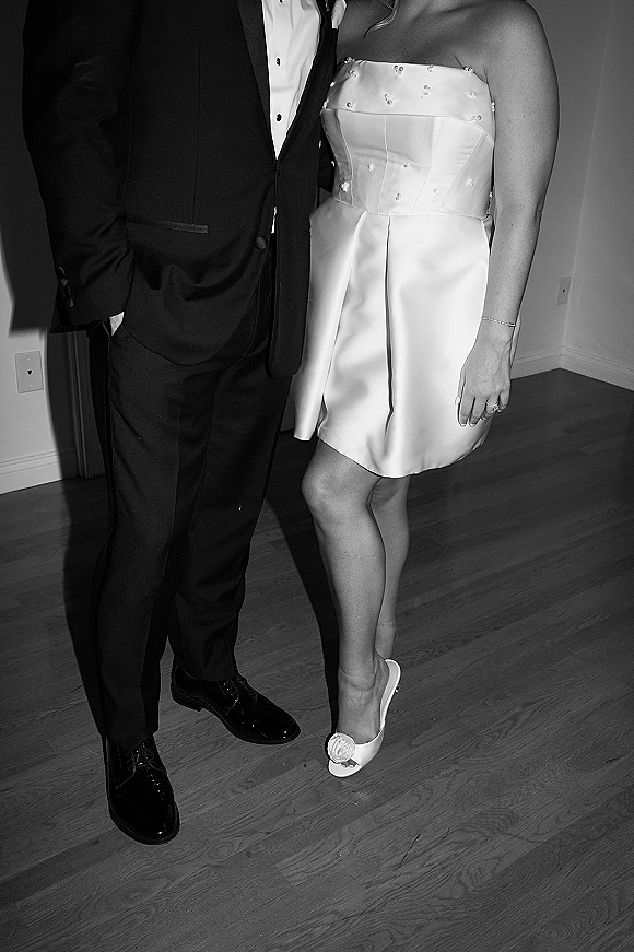 Couple portrait in a black and white wedding portrait, bride in strapless satin mini dress with pearls beside groom in tuxedo indoors