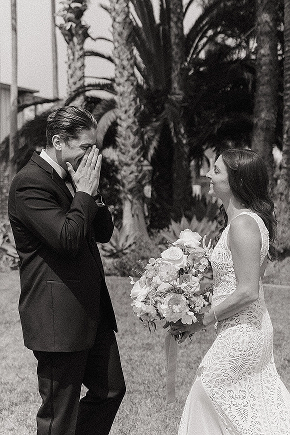 Wedding first look as bride holds a white rose bouquet and smiles at groom in tuxedo covering his face on a palm-lined lawn