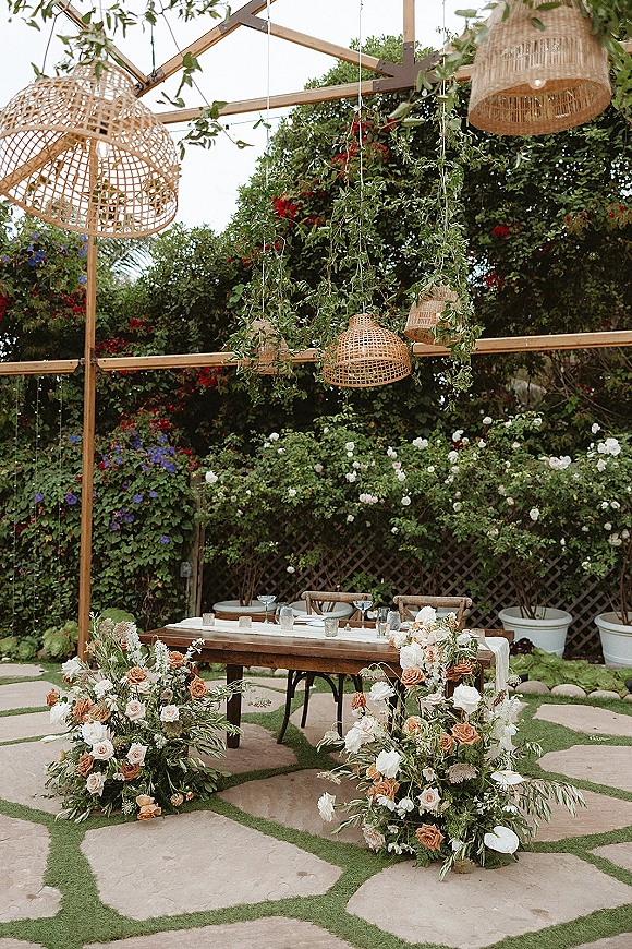 Reception tablescape on a wood farmhouse outdoor reception table with white runner, rose greenery garlands, and rattan pendant lanterns by a lattice fence