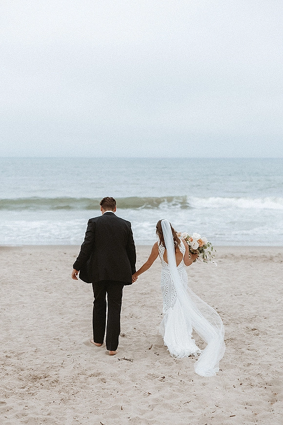 Couple portrait of beach wedding couple walking away hand in hand, bride in wedding dress with long veil and bouquet on sandy shore by ocean waves