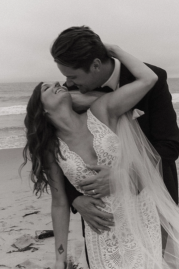 Wedding couple portrait in black and white, groom in tuxedo dipping and kissing bride in lace dress with veil by ocean waves