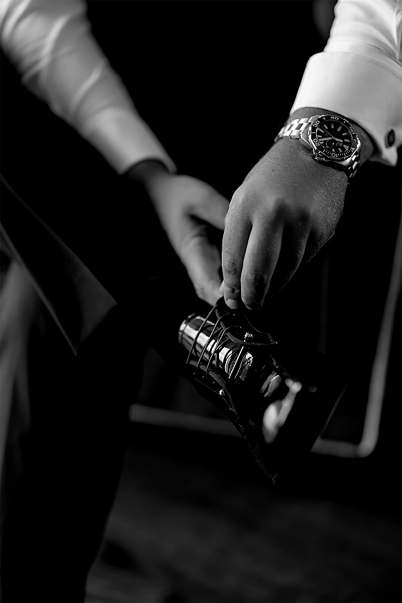 Groom getting ready, tying black leather dress shoes with wristwatch accent, in a dark indoor room with a chair nearby