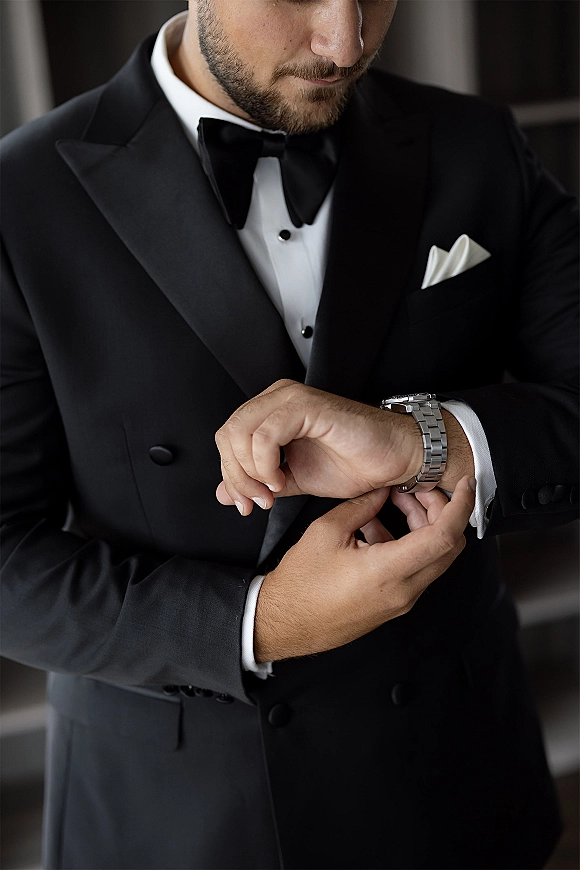 Groom getting ready, adjusting watch with cufflinks and pocket square on a black tuxedo in soft window light indoors