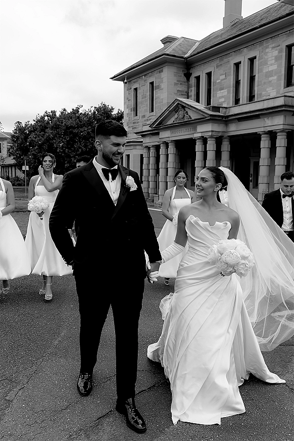 Wedding party portrait of bride in veil and gloves with bouquet beside groom in black tux, walking past a stone building with columns