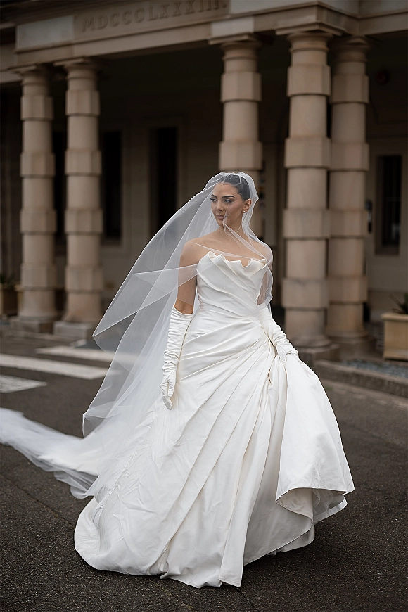 Bridal portrait of a bride in an off the shoulder wedding dress with cathedral veil and opera gloves on stone steps by columns