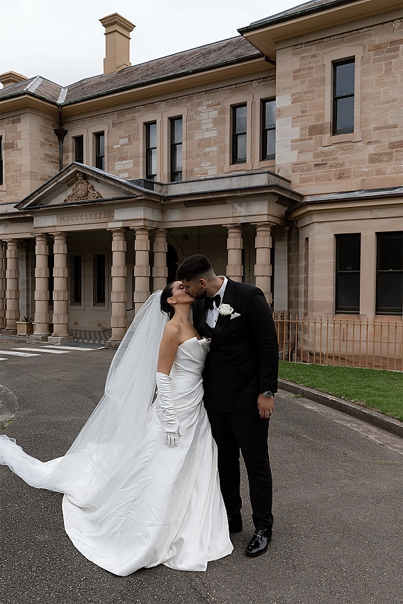 Wedding kiss portrait of bride and groom kissing, her cathedral veil flowing over a strapless gown and gloves, by a stone columned building